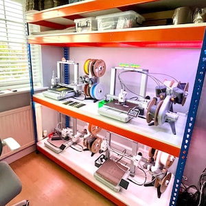 May include: A multi-tiered storage shelf with orange and blue accents, filled with 3D printers and spools of filament. The printers are silver and black, with various components and wires. The shelf is in a well-lit room, suggesting a workshop or hobby space.