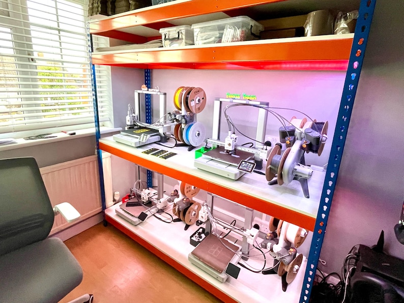 May include: A multi-tiered storage shelf with orange and blue accents, filled with 3D printers and spools of filament. The printers are silver and black, with various components and wires. The shelf is in a well-lit room, suggesting a workshop or hobby space.