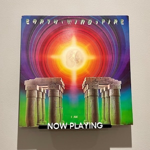 May include: A vinyl record album cover featuring a colorful, psychedelic design with a bright yellow sun and pillars. The album is titled "Earth, Wind & Fire" and the text "I Am" is visible at the bottom. The text "NOW PLAYING" is displayed in black at the bottom of the image.