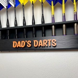 May include: A black dart holder with the text "DAD'S DARTS" in orange. The holder displays several darts with yellow, blue, and white flights. The darts are arranged neatly, showcasing their colorful design and professional appearance.