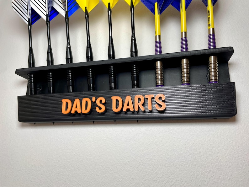 May include: A black dart holder with the text "DAD'S DARTS" in orange. The holder displays several darts with yellow, blue, and white flights. The darts are arranged neatly, showcasing their colorful design and professional appearance.