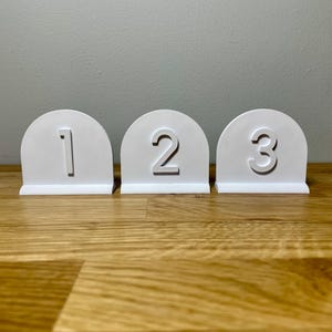 Wedding table numbers in white standing up on a wooden desk with grey background. Numbers 1 2 3 shown