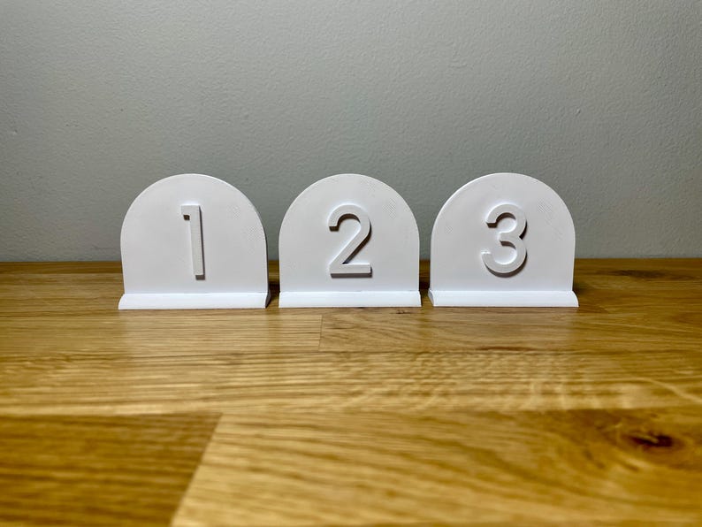 Wedding table numbers in white standing up on a wooden desk with grey background. Numbers 1 2 3 shown