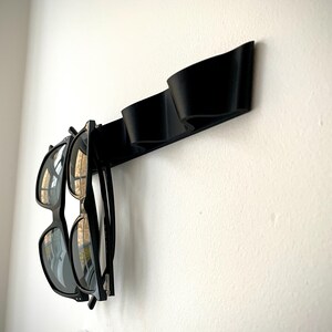 May include: A black wall-mounted sunglasses holder with two pairs of sunglasses hanging from it. The holder is made of a curved, solid material and is mounted on a white wall. The sunglasses have black frames and dark lenses.