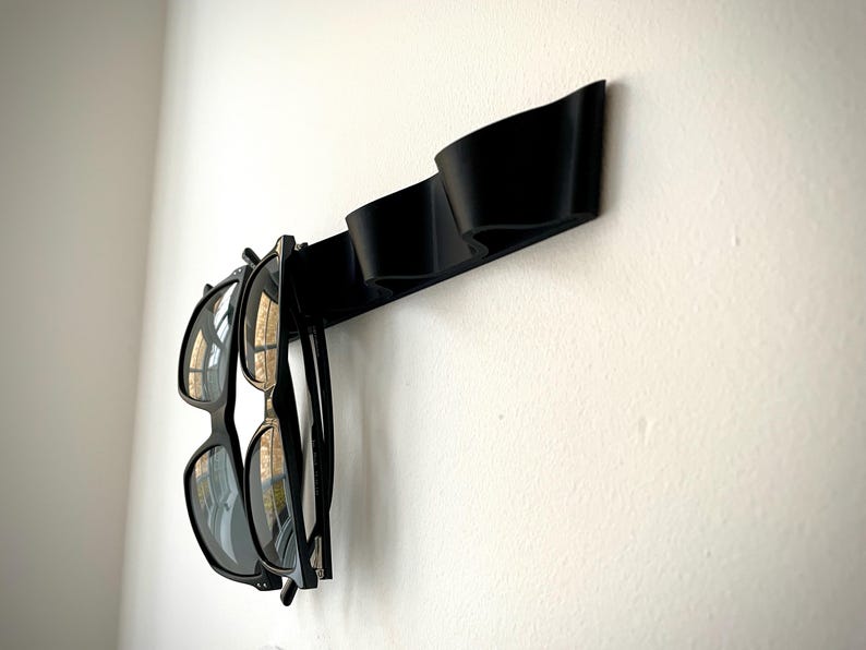 May include: A black wall-mounted sunglasses holder with two pairs of sunglasses hanging from it. The holder is made of a curved, solid material and is mounted on a white wall. The sunglasses have black frames and dark lenses.
