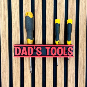 May include: A black and red "DAD'S TOOLS" sign with three screwdrivers. The sign is mounted on a wooden wall with vertical stripes. The screwdrivers have yellow and black handles.
