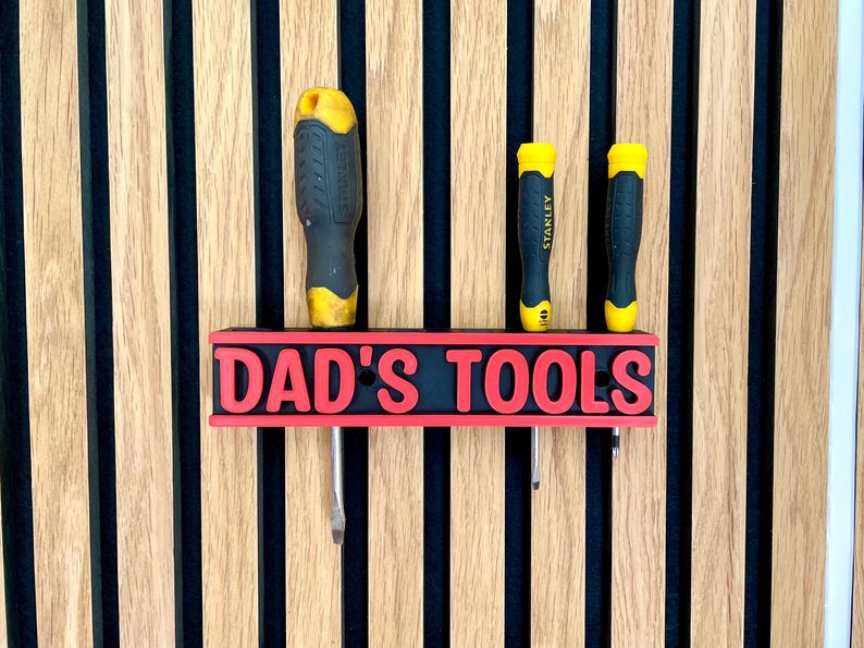 May include: A black and red "DAD'S TOOLS" sign with three screwdrivers. The sign is mounted on a wooden wall with vertical stripes. The screwdrivers have yellow and black handles.