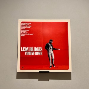 May include: A red vinyl record album cover featuring a black and white photograph of a man wearing a jacket and trousers. The album is titled "Leon Bridges Coming Home" and lists the songs "Coming Home", "Better Man", "Brown Skin Girl", "Smooth Sailin", "Shine", "Lisa Sawyer", "Flowers", "Pull Away", "Twistin' & Groovin", and "River".