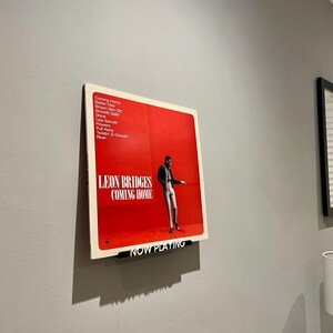 May include: A red vinyl record with a black and white photo of a man in a suit. The record is titled "Leon Bridges Coming Home" and has the text "Now Playing" on a black bar at the bottom.
