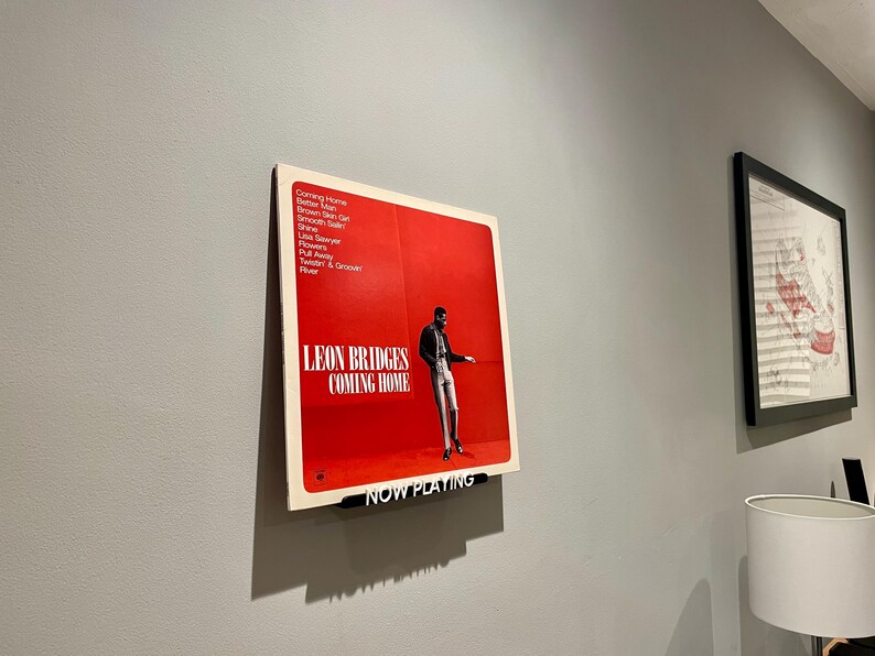 May include: A red vinyl record with a black and white photo of a man in a suit. The record is titled "Leon Bridges Coming Home" and has the text "Now Playing" on a black bar at the bottom.