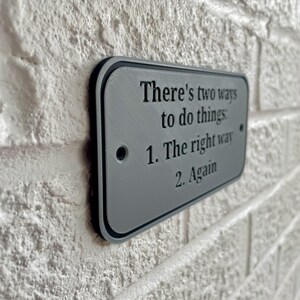 May include: A rectangular gray sign with black trim is mounted on a white brick wall. The sign reads, "There's two ways to do things: 1. The right way 2. Again."