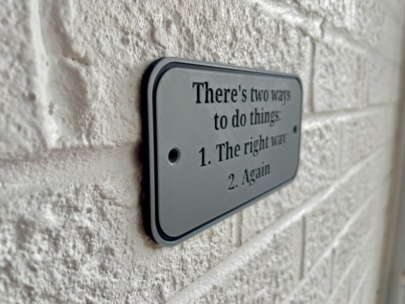 May include: A rectangular gray sign with black trim is mounted on a white brick wall. The sign reads, "There's two ways to do things: 1. The right way 2. Again."