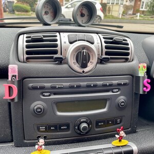 Pu&ograve; includere: Il cruscotto di un'auto con una radio nera, bocchette di ventilazione e una lettera D e S nera e rosa sul lato. Ci sono due figurine di Topolino sul cruscotto.