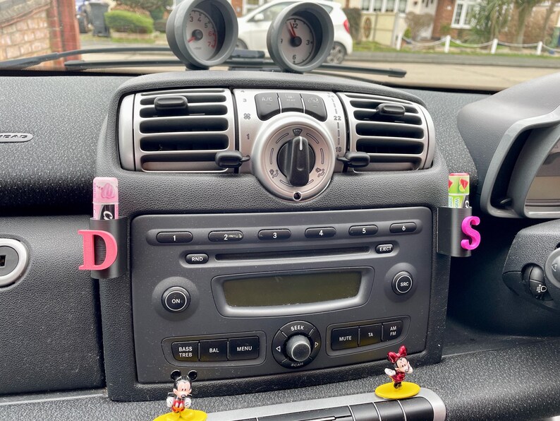 Pu&ograve; includere: Il cruscotto di un'auto con una radio nera, bocchette di ventilazione e una lettera D e S nera e rosa sul lato. Ci sono due figurine di Topolino sul cruscotto.