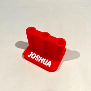 Op de afbeelding: Een helderrode 3D-geprinte bureau-organizer met drie ronde gaten. De naam "JOSHUA" is in het wit op de basis gedrukt. Ontworpen om pennen, potloden of andere kleine kantoorbenodigdheden in op te bergen.