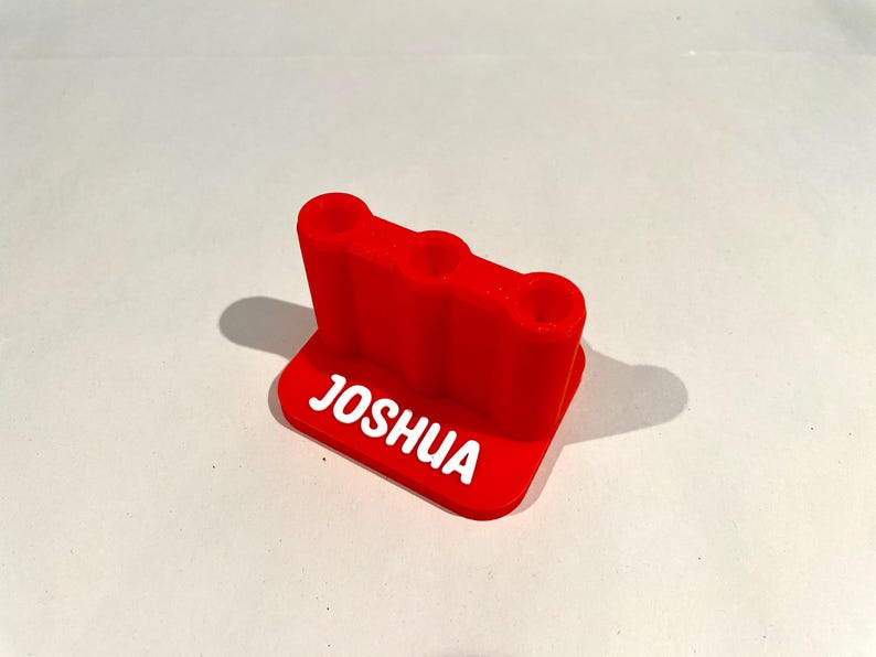 Op de afbeelding: Een helderrode 3D-geprinte bureau-organizer met drie ronde gaten. De naam "JOSHUA" is in het wit op de basis gedrukt. Ontworpen om pennen, potloden of andere kleine kantoorbenodigdheden in op te bergen.