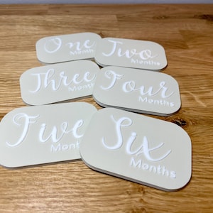May include: Six beige oval-shaped baby milestone cards with white text. The cards are numbered one through six and say "Month" or "Months".