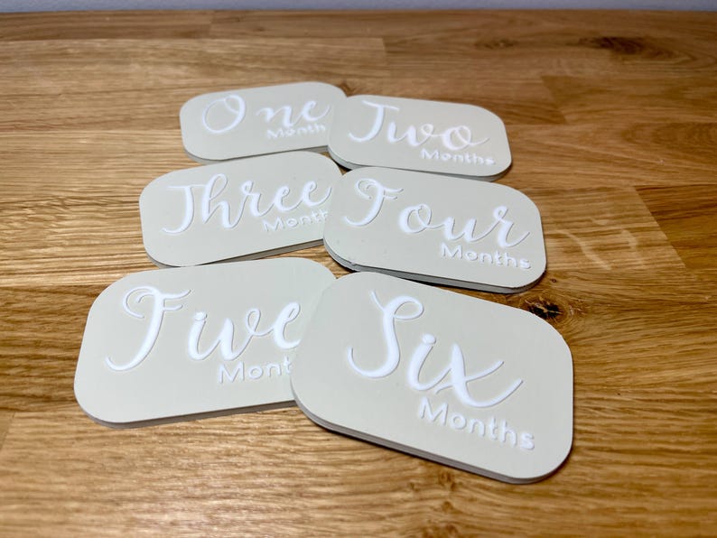 May include: Six beige oval-shaped baby milestone cards with white text. The cards are numbered one through six and say "Month" or "Months".