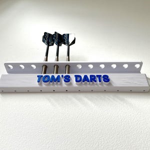 May include: A white dart holder with the text "TOM'S DARTS" in blue. Three darts with black and white flights are inserted. The holder is mounted on a white wall.