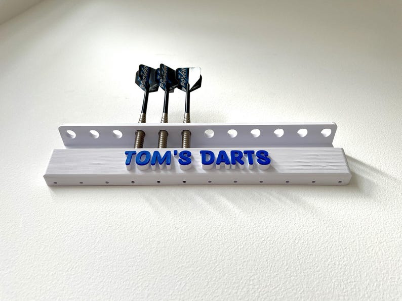 May include: A white dart holder with the text "TOM'S DARTS" in blue. Three darts with black and white flights are inserted. The holder is mounted on a white wall.