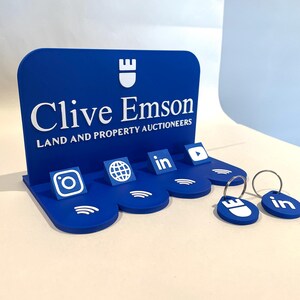 May include: A blue display stand with the text "Clive Emson" and social media icons. The stand has a curved top and a base with slots for keychains. Two blue keychains with silver rings are in the foreground.