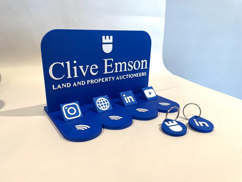 May include: A blue display stand with the text "Clive Emson" and social media icons. The stand has a curved top and a base with slots for keychains. Two blue keychains with silver rings are in the foreground.