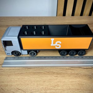 Pu&ograve; includere: Un organizer da scrivania a forma di camion grigio e arancione. Il camion ha una cabina grigia e un'area di carico arancione con le lettere bianche "LS". L'organizer ha scomparti per penne e altri accessori. Il camion &egrave; lungo circa 25 cm.