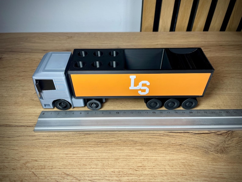 Pu&ograve; includere: Un organizer da scrivania a forma di camion grigio e arancione. Il camion ha una cabina grigia e un'area di carico arancione con le lettere bianche "LS". L'organizer ha scomparti per penne e altri accessori. Il camion &egrave; lungo circa 25 cm.