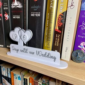 May include: A white wooden sign with the text "Days until our Wedding" and two gray hearts with the numbers 9 and 6 on them. The sign is sitting on a bookshelf with a variety of books.