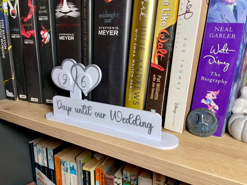 May include: A white wooden sign with the text "Days until our Wedding" and two gray hearts with the numbers 9 and 6 on them. The sign is sitting on a bookshelf with a variety of books.