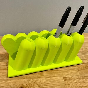 May include: A bright neon green 3D-printed desk organizer featuring a series of cylindrical pen holders arranged in a row, with a heart-shaped accent on one end. The organizer holds two grey markers and one black pen, all standing upright in the slots.