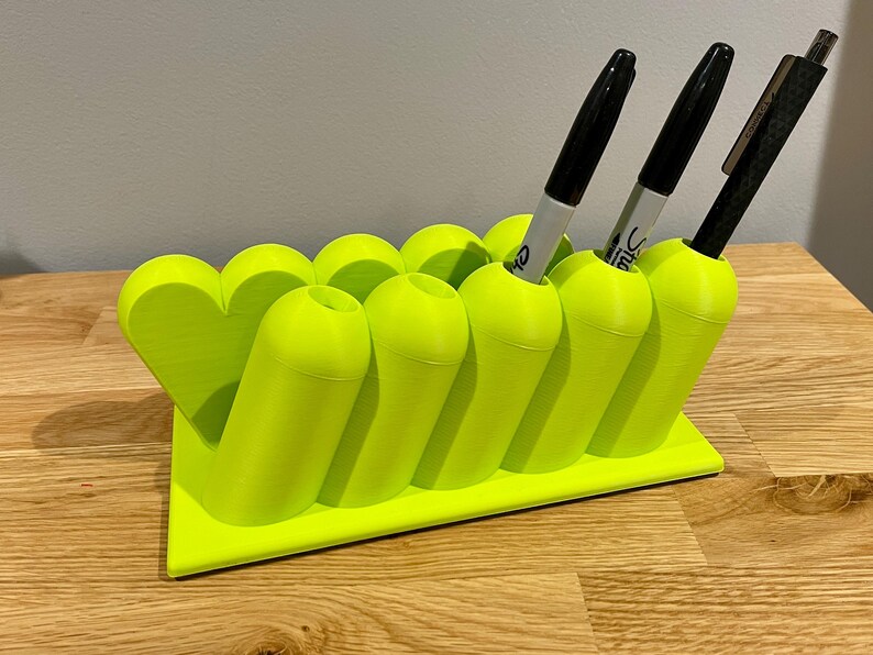 May include: A bright neon green 3D-printed desk organizer featuring a series of cylindrical pen holders arranged in a row, with a heart-shaped accent on one end. The organizer holds two grey markers and one black pen, all standing upright in the slots.