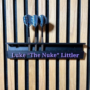 Puede incluir: Soporte negro para dardos con tres dardos. El soporte tiene el nombre "Luke 'The Nuke' Littler" en letras moradas. Los dardos tienen ca&ntilde;as negras y plumas Power azules y negras. El fondo es una pared de madera.