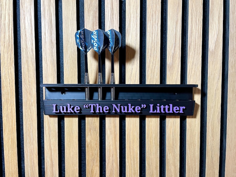 Puede incluir: Soporte negro para dardos con tres dardos. El soporte tiene el nombre "Luke 'The Nuke' Littler" en letras moradas. Los dardos tienen ca&ntilde;as negras y plumas Power azules y negras. El fondo es una pared de madera.