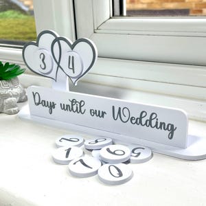 May include: White wooden countdown calendar with two hearts and the text "Days until our Wedding". The calendar has flippable numbers and includes a set of numbers 0-9.
