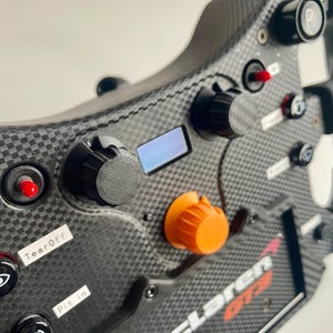 May include: Close-up of a black racing steering wheel with a carbon fiber finish. The wheel features multiple buttons and knobs, including a red button labeled "TC", a red button labeled "TearOff", a black knob, and an orange knob. The text "McLaren GT3" is visible on the bottom right of the wheel.