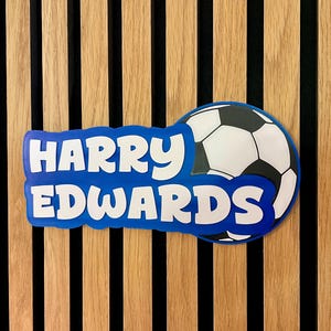 May include: A blue and white soccer-themed nameplate with the name "HARRY EDWARDS" in white letters. The nameplate is in front of a soccer ball graphic. The background is a wooden wall with vertical wood panels.