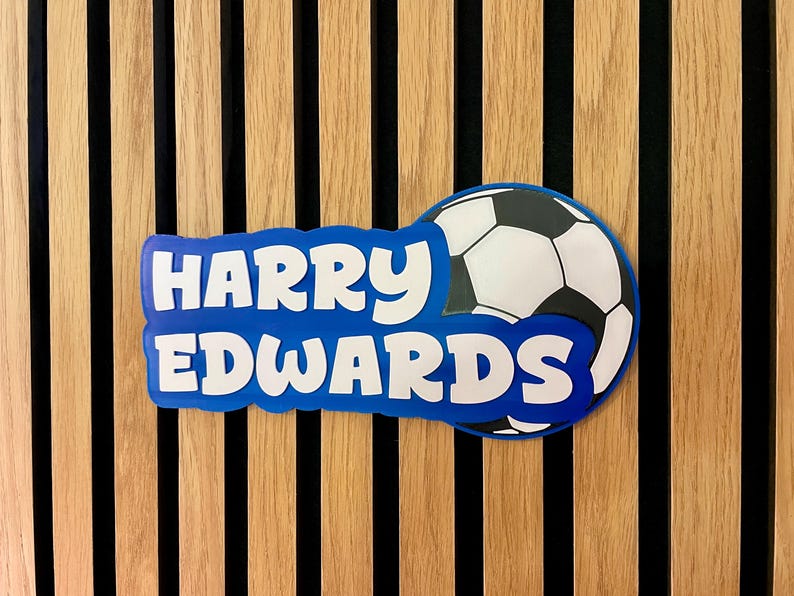 May include: A blue and white soccer-themed nameplate with the name "HARRY EDWARDS" in white letters. The nameplate is in front of a soccer ball graphic. The background is a wooden wall with vertical wood panels.