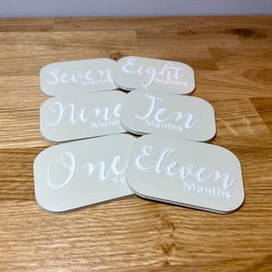May include: Set of six beige baby milestone cards with white text. The cards feature the numbers one through ten, and eleven, with the word "months" written after each number. The cards are arranged in a semi-circle.