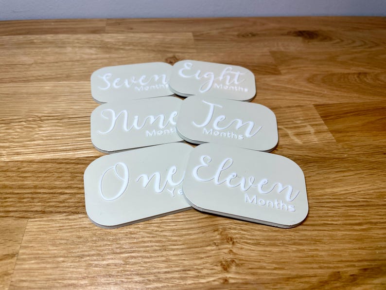 May include: Set of six beige baby milestone cards with white text. The cards feature the numbers one through ten, and eleven, with the word "months" written after each number. The cards are arranged in a semi-circle.