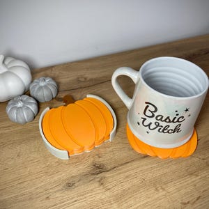 Puede incluir: Una taza de cer&aacute;mica blanca con el texto "Basic Witch" y un posavasos con forma de calabaza. La taza est&aacute; sobre un posavasos naranja. Tambi&eacute;n hay calabazas decorativas blancas y grises.