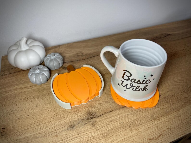 Puede incluir: Una taza de cer&aacute;mica blanca con el texto "Basic Witch" y un posavasos con forma de calabaza. La taza est&aacute; sobre un posavasos naranja. Tambi&eacute;n hay calabazas decorativas blancas y grises.