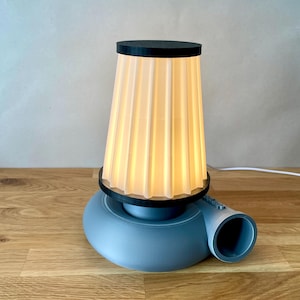 Puede incluir: Una l&aacute;mpara de mesa azul y negra con una pantalla blanca plisada. La l&aacute;mpara tiene una base &uacute;nica que se asemeja a un ventilador.