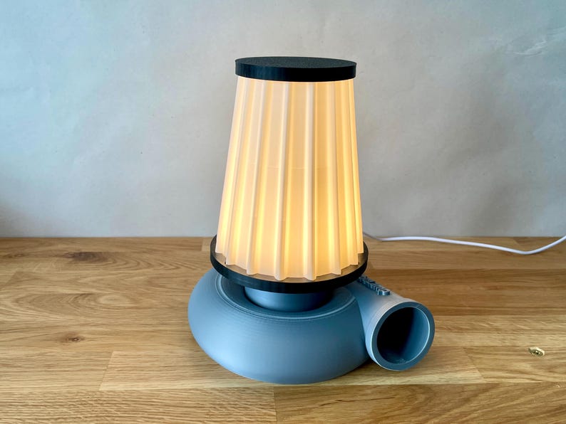 Puede incluir: Una l&aacute;mpara de mesa azul y negra con una pantalla blanca plisada. La l&aacute;mpara tiene una base &uacute;nica que se asemeja a un ventilador.
