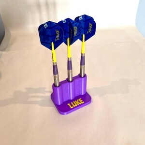 Op de afbeelding: Drie darts met blauwe en gele flights in een paarse houder. De darts hebben zilveren schachten en gele punten. De houder is gepersonaliseerd met de naam "LUKE" in geel.