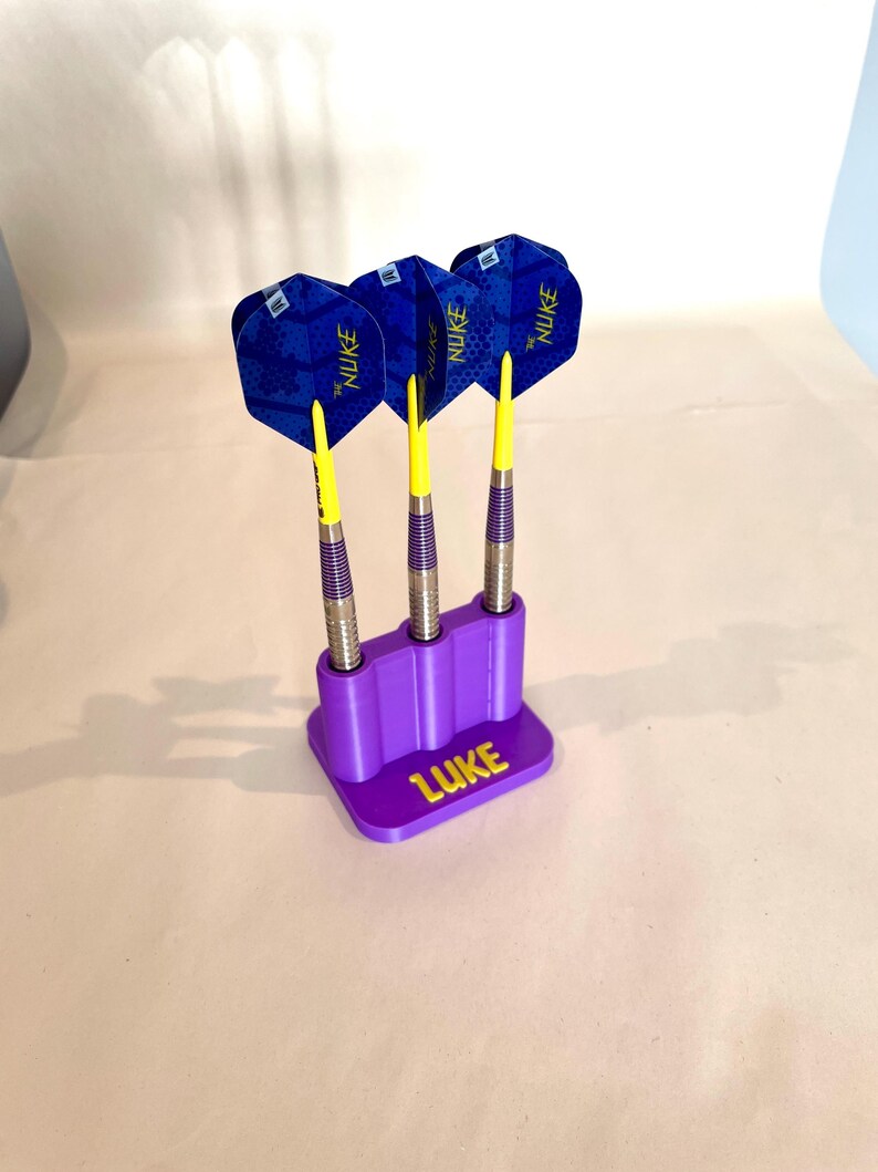 Op de afbeelding: Drie darts met blauwe en gele flights in een paarse houder. De darts hebben zilveren schachten en gele punten. De houder is gepersonaliseerd met de naam "LUKE" in geel.