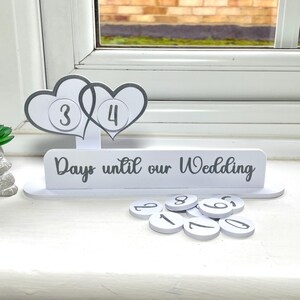 May include: White wooden countdown calendar with two hearts and the text "Days until our Wedding". The hearts have the numbers 3 and 4 on them. There are also several white circles with numbers on them.