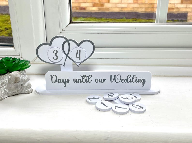 May include: White wooden countdown calendar with two hearts and the text "Days until our Wedding". The hearts have the numbers 3 and 4 on them. There are also several white circles with numbers on them.