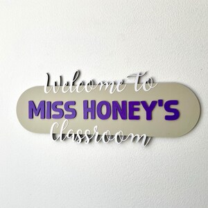 K&ouml;nnte beinhalten: Ein Klassenzimmerschild mit dem Text "Welcome to MISS HONEY'S Classroom". Das Schild hat einen beigen ovalen Hintergrund mit lila Buchstaben und wei&szlig;em Schreibschrifttext. Es ist an einer wei&szlig;en Wand befestigt.