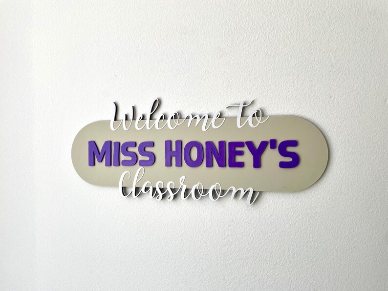 K&ouml;nnte beinhalten: Ein Klassenzimmerschild mit dem Text "Welcome to MISS HONEY'S Classroom". Das Schild hat einen beigen ovalen Hintergrund mit lila Buchstaben und wei&szlig;em Schreibschrifttext. Es ist an einer wei&szlig;en Wand befestigt.